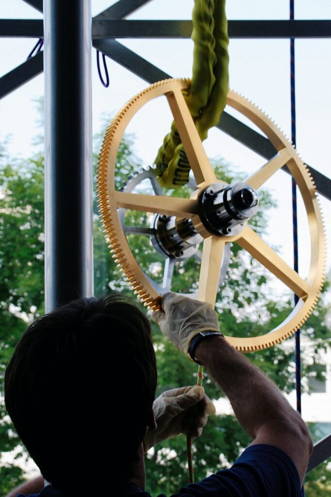 Ein großes Zahnrad der Turmuhr Monumentum Temporis wird bei der Montage an Seilen nach oben gezogen.