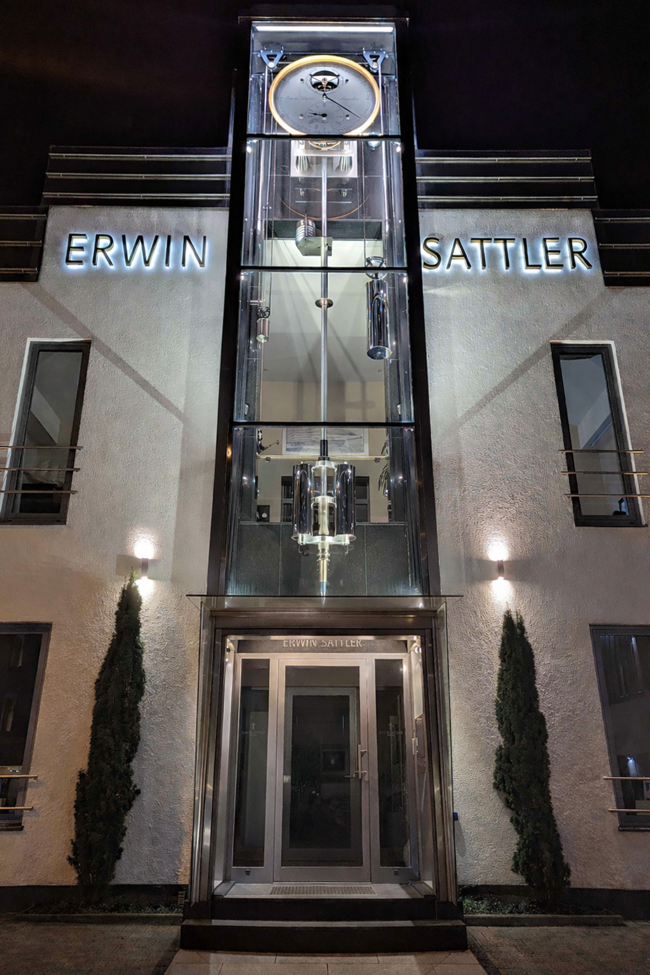 Das beleuchtete Manufakturgebäude von Erwin Sattler bei Nacht mit der fertigen Turmuhr Monumentum Temporis davor.
